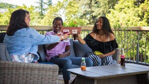 Trois personnes trinquent avec des boissons sur une terrasse aménagée avec des meubles en osier et une table, le tout entouré de verdure.