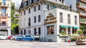 Il s’agit d’un bâtiment historique sur lequel on peut voir une enseigne indiquant le restaurant de l’hôtel des Bains. Il est doté de nombreuses fenêtres, d’un balcon et offre une vue sur la rue, où l’on aperçoit des voitures garées.