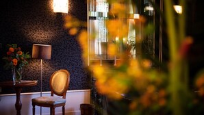 Une pièce avec un mur orné d’un motif, une chaise en bois, une petite table sur laquelle est posé un vase de fleurs et un lampadaire.