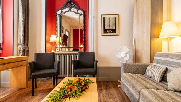 Une chambre d’hôtel avec un bureau en bois, deux fauteuils, un canapé, un miroir, un ventilateur et un arrangement floral placé sur la table basse.