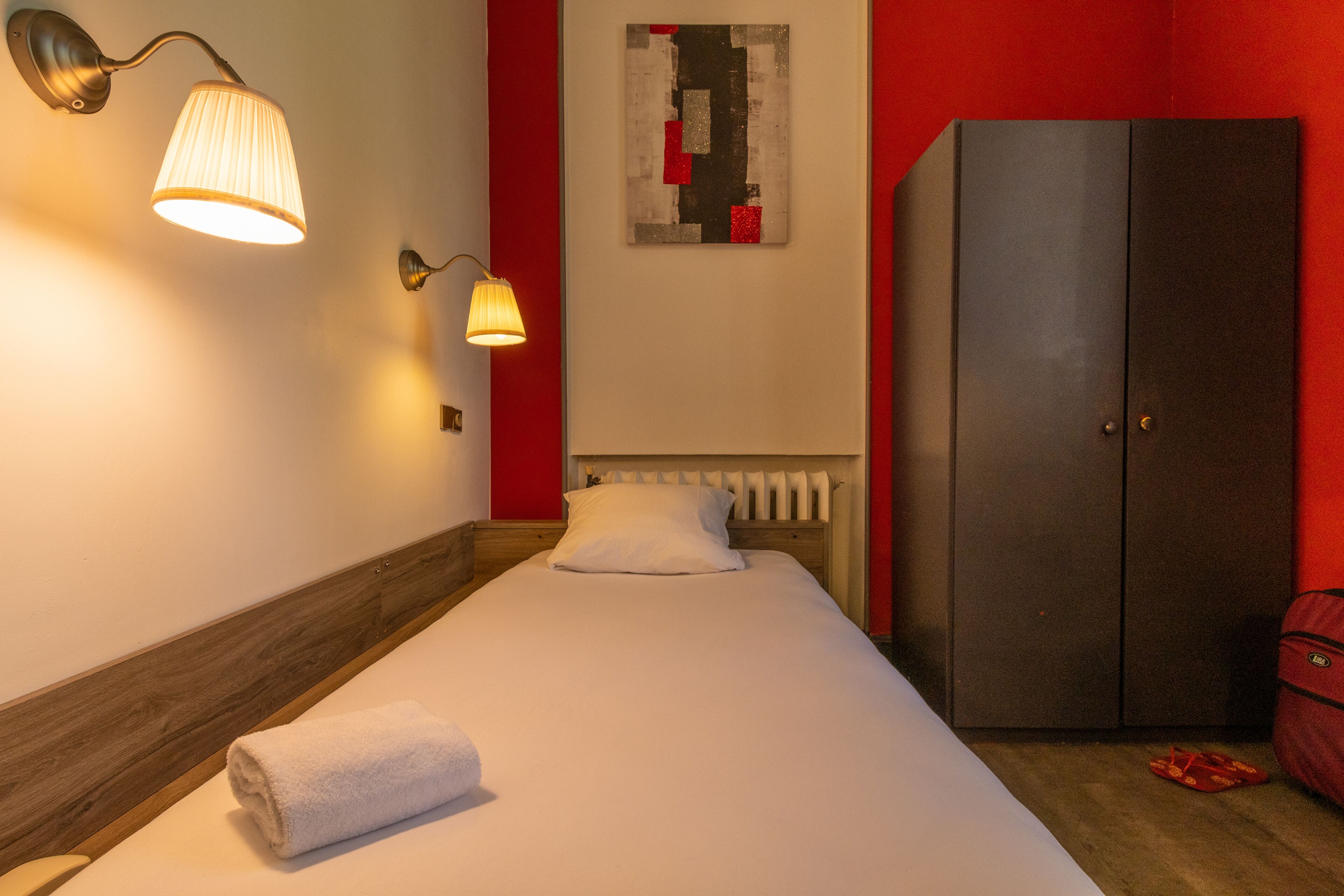 A neatly made bed with a white blanket and a rolled towel, a wooden headboard, a wall-mounted lamp, a red cabinet, and a piece of abstract art on the wall.