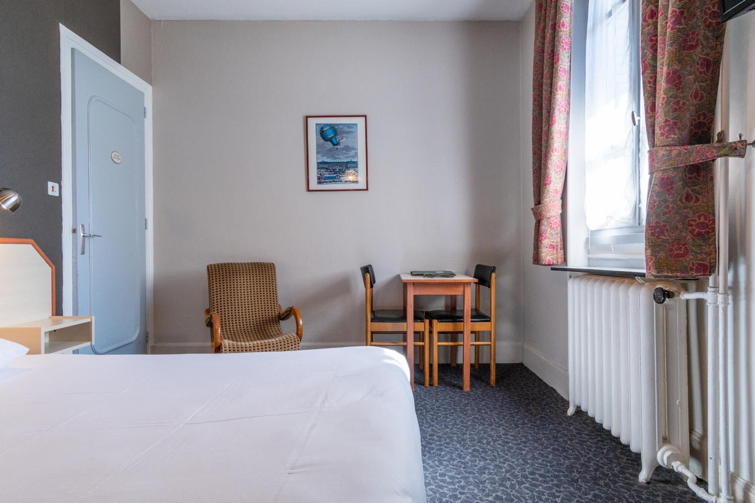 A hotel room with a bed, a chair, a table, a framed picture on the wall, and a window with curtains.