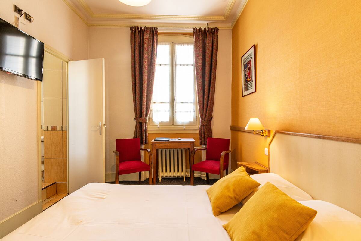 Een hotelkamer met een bed, twee rode stoelen, een klein tafeltje, een radiator, een aan de muur bevestigde televisie en een raam met gordijnen.