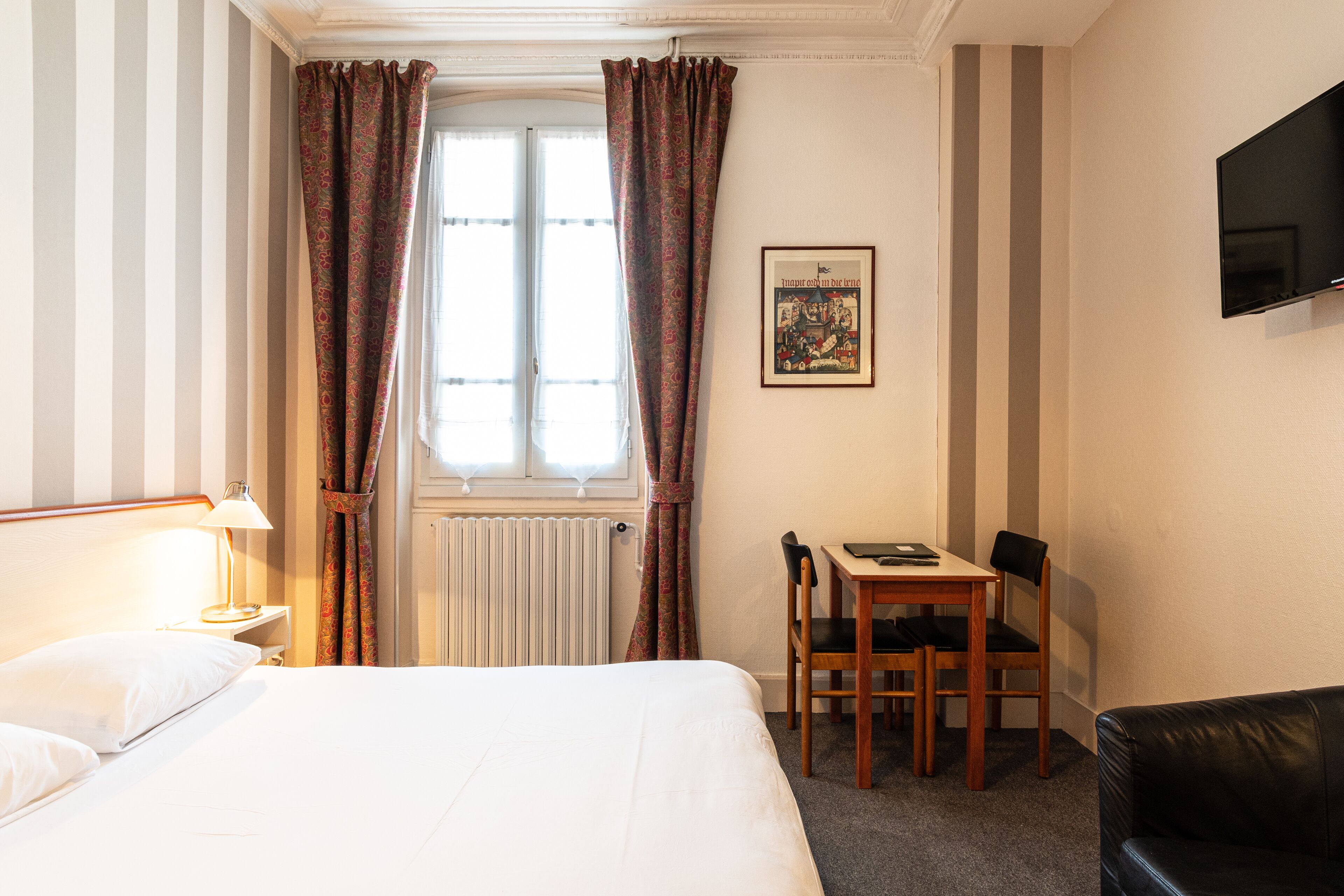 Habitación de hotel con cama, escritorio con silla, televisor montado en la pared y ventana con cortinas.