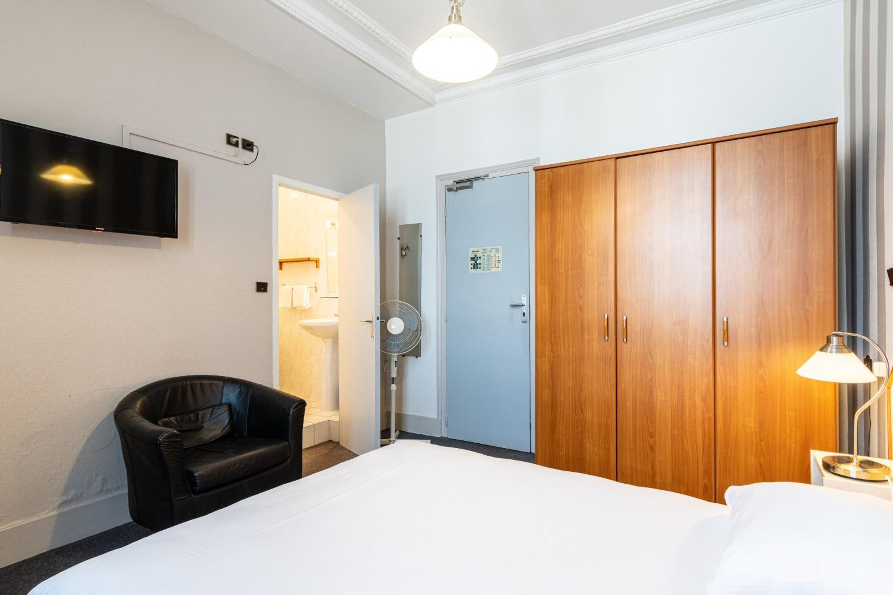 A hotel room with a bed, a black leather chair, a wooden wardrobe, a television mounted on the wall, and a fan.