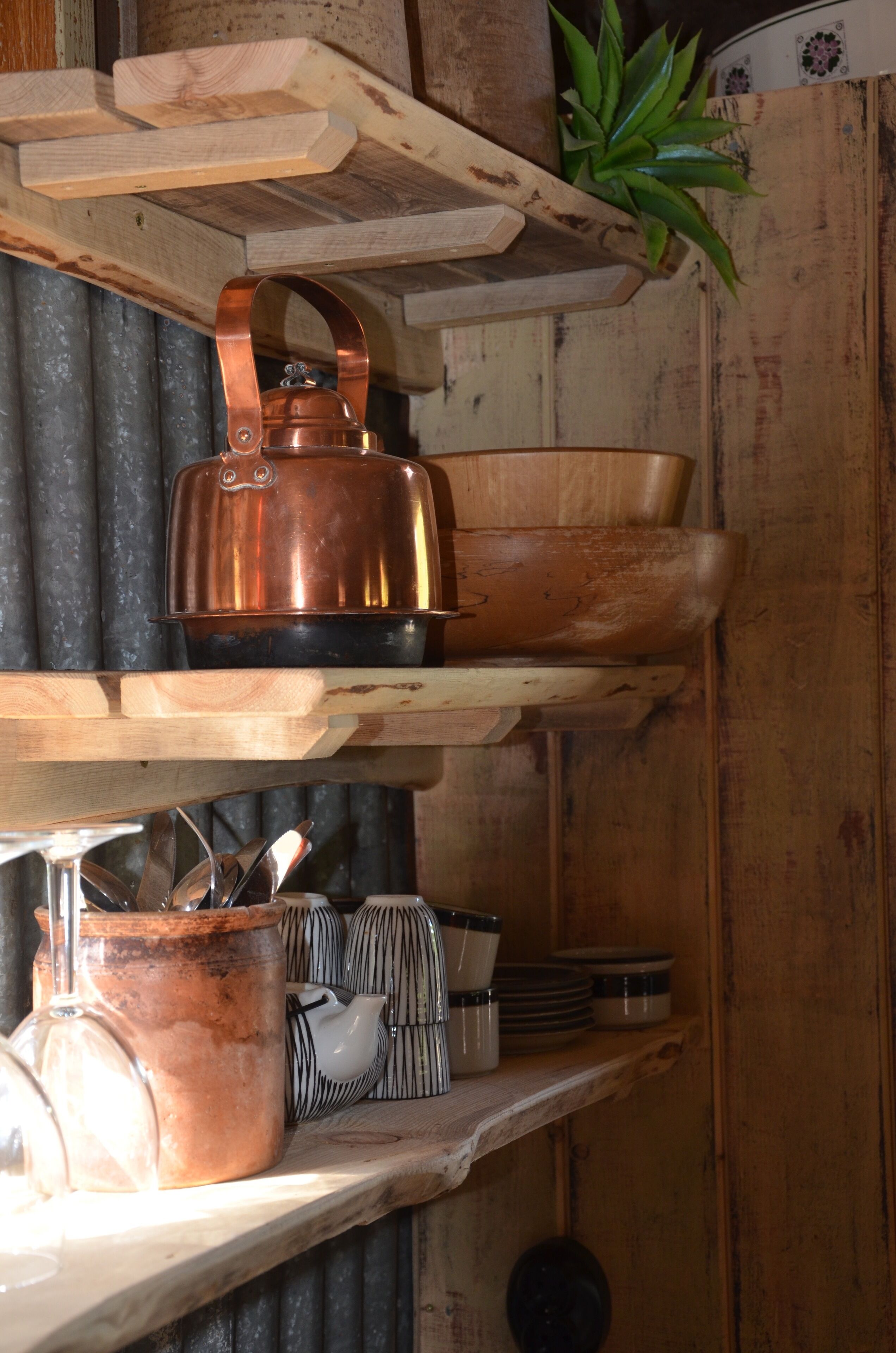 A rustic kitchen with wooden shelves holding copper cookware, a kettle, and striped mugs.
