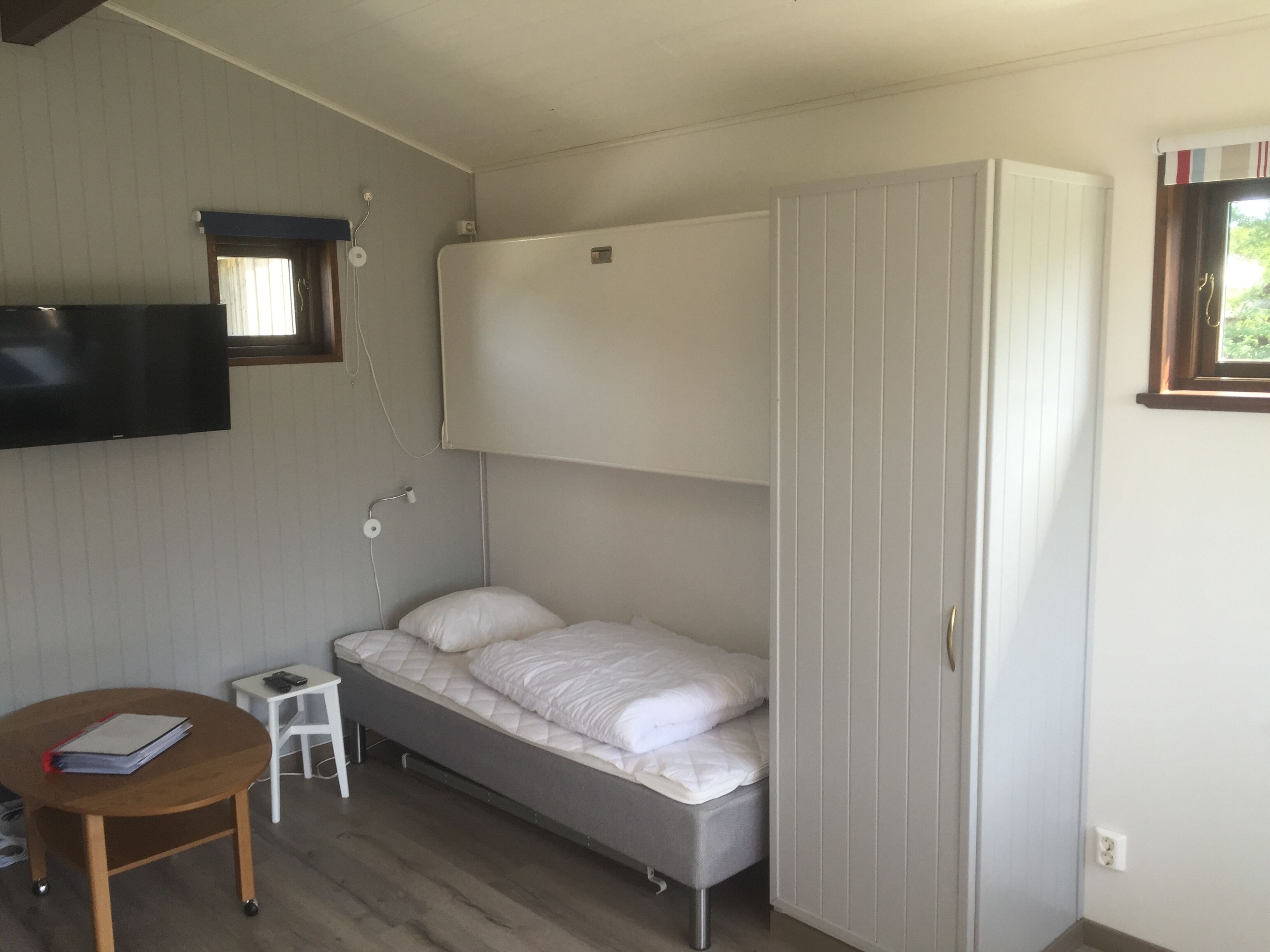 Ein kleines Zimmer mit einem Bett, einem Tisch, einem an der Wand befestigten Fernseher und einem Fenster mit Blick auf Grünfläche.