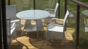 Eine Terrasse mit Glasgeländer, die auf eine Wiese und ein Gewässer blickt.