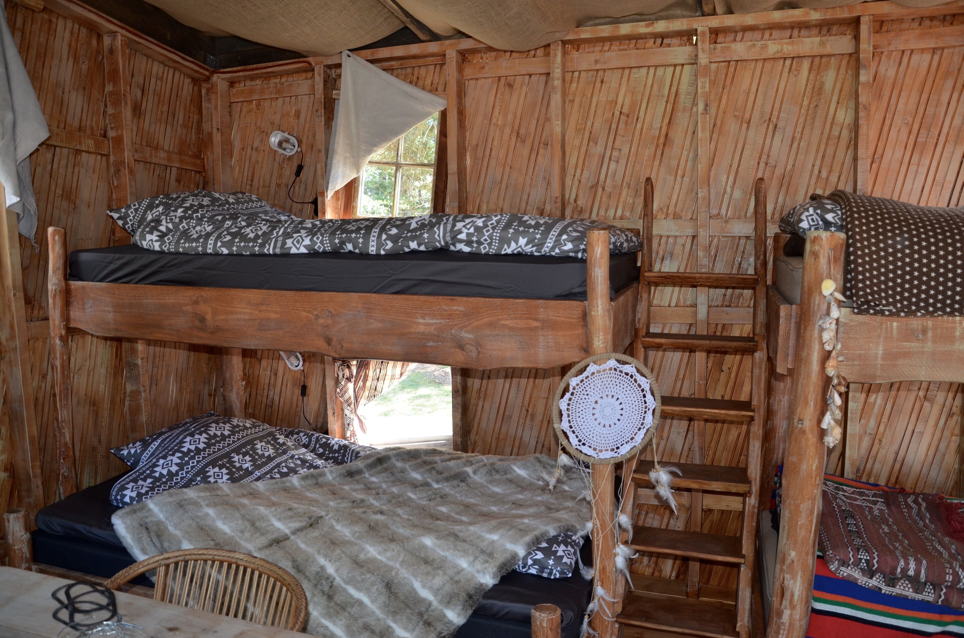 Una habitación rústica de madera con una cama litera, una cama individual, una silla de mimbre y una mesita pequeña.