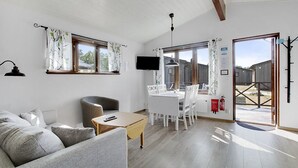 Ein modernes Wohnzimmer mit einer Couch, einem kleinen Tisch und einem Essbereich mit weißem Tisch und Stühlen. An der Wand ist ein Fernseher befestigt und ein Feuerlöscher hängt dort.