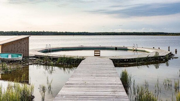 Eine Holzstele führt zu einem runden Pool mit einer Sitzbank in der Mitte, umgeben von ruhigem Wasser und einer fernen Küstenlinie.