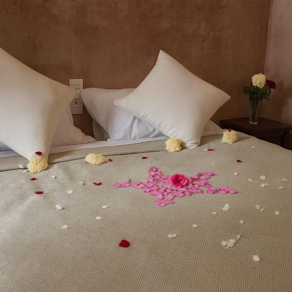 Un lit bien fait, avec des oreillers blancs et une couvre-lit beige, agrémenté de p étales de rose éparpillés et d’une seule rose rose.