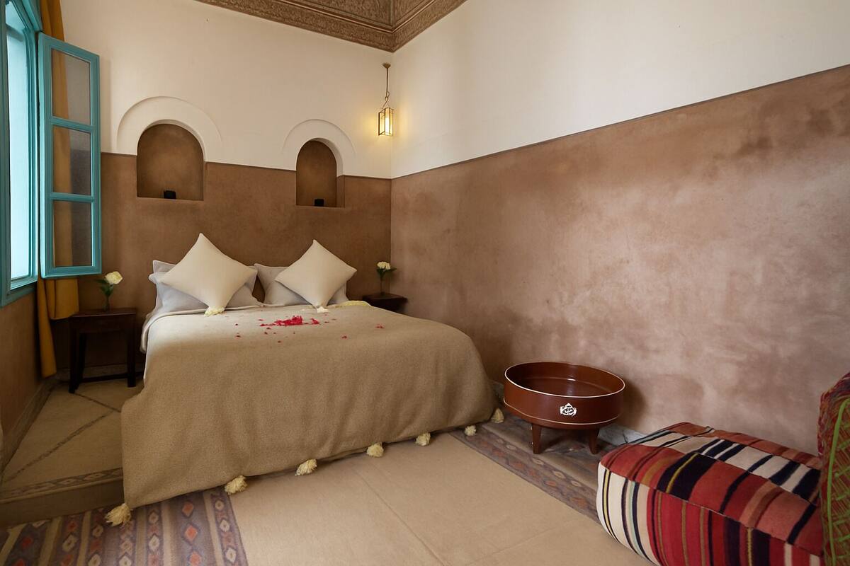 Un dormitorio con cama, una mesita de noche, una lámpara de pared, una pequeña mesa con un florero y un cojín colorido.