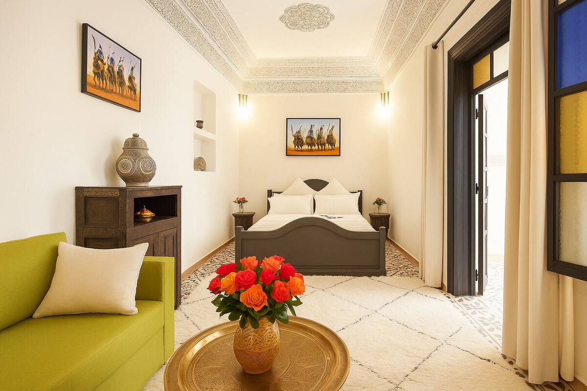 Habitación de hotel con cama, sofá verde, un jarrón decorativo con flores y dos cuadros en la pared.
