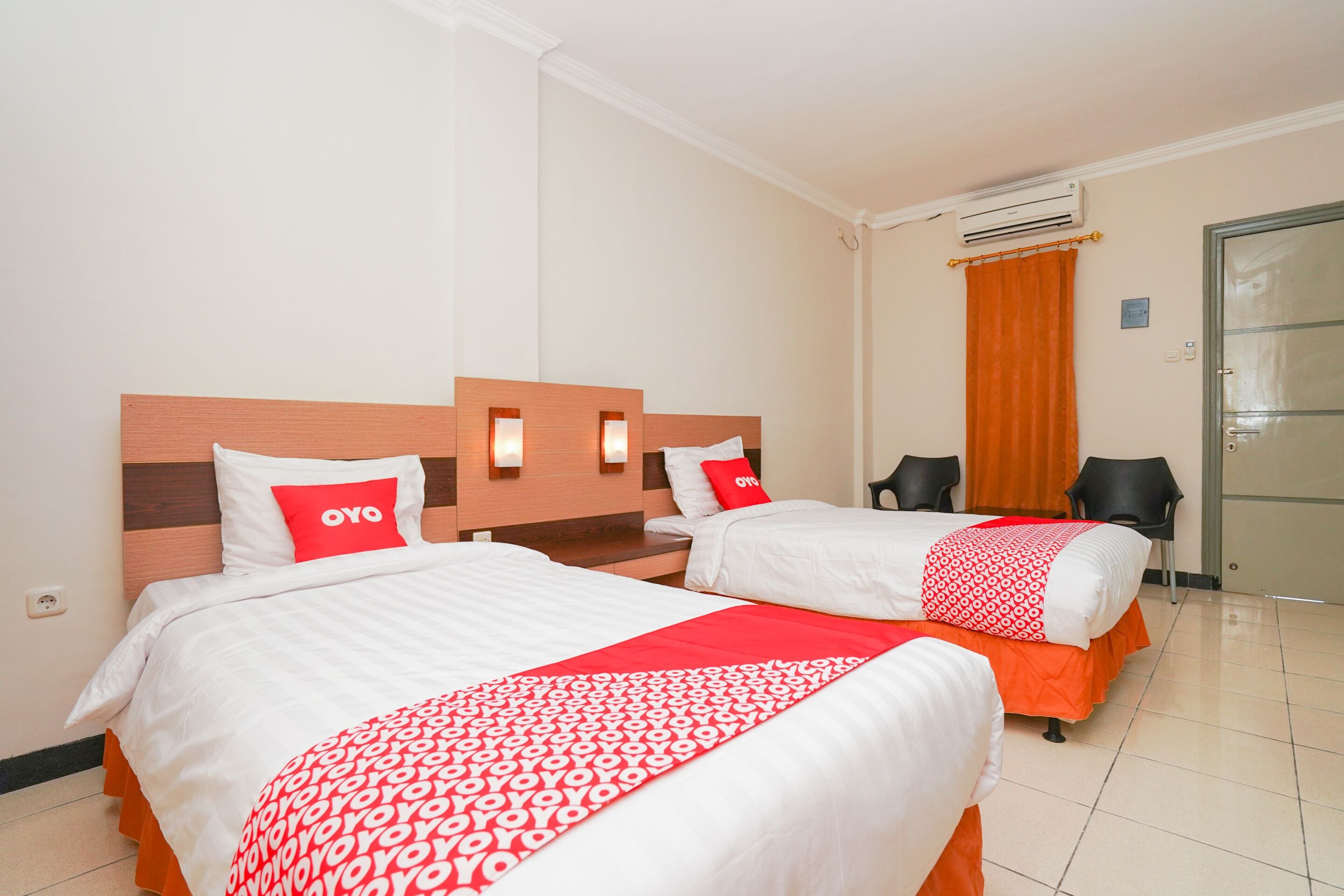 A hotel room with two beds, each with white bedding and red accents, a wooden headboard, and an air conditioning unit on the wall.