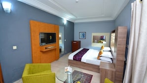 Une chambre d’hôtel avec un lit, un téléviseur fixé sur un panneau en bois, une table basse en verre et deux fauteuils jaunes.