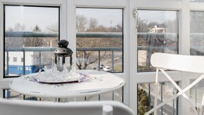 Una mesa con mantel floral, una vela y copas, frente a una ventana que ofrece vistas a edificios y árboles.
