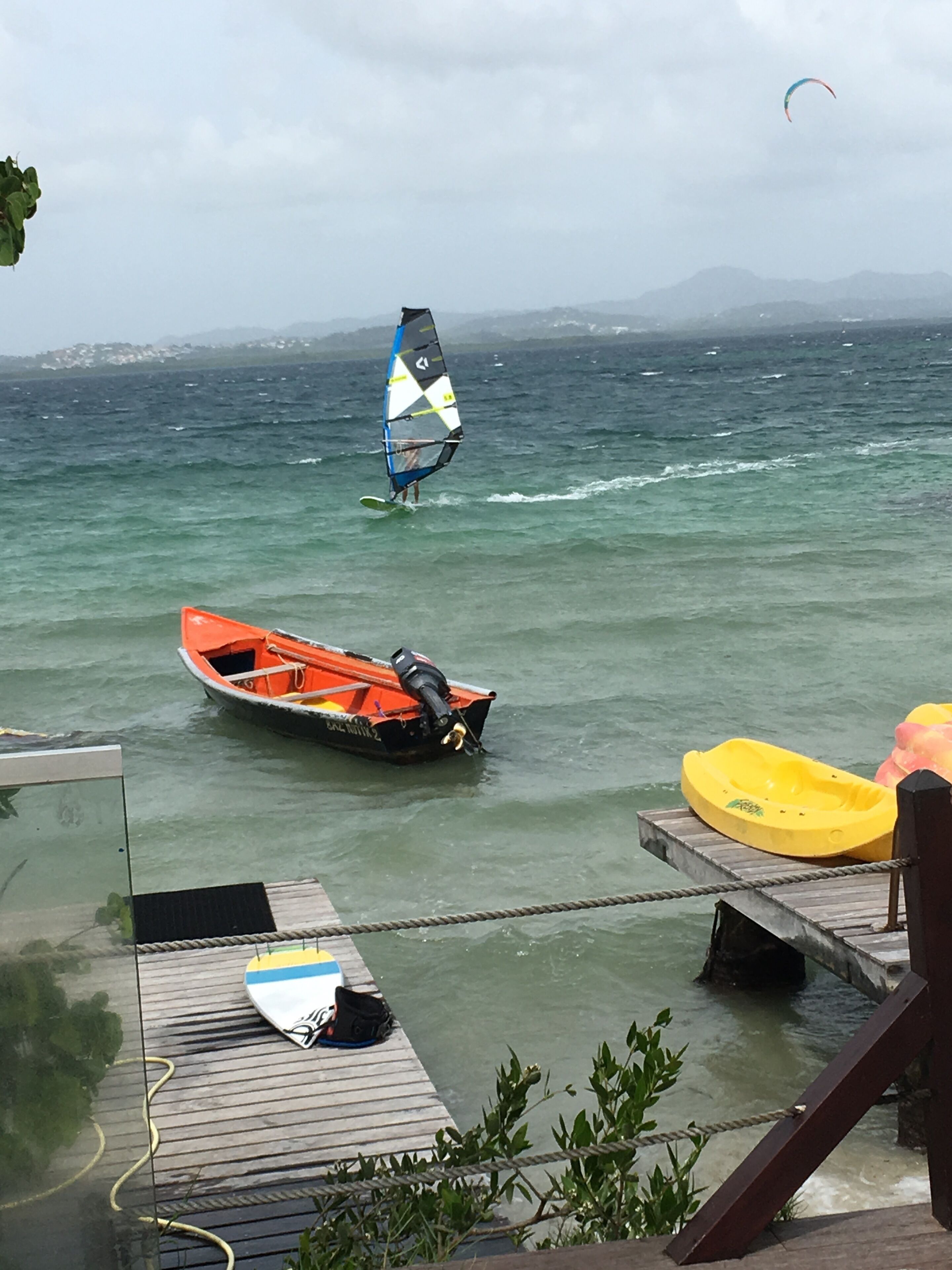 A wooden dock with a surfboard and a kayak, a small orange boat, and a sailboarder in the water.