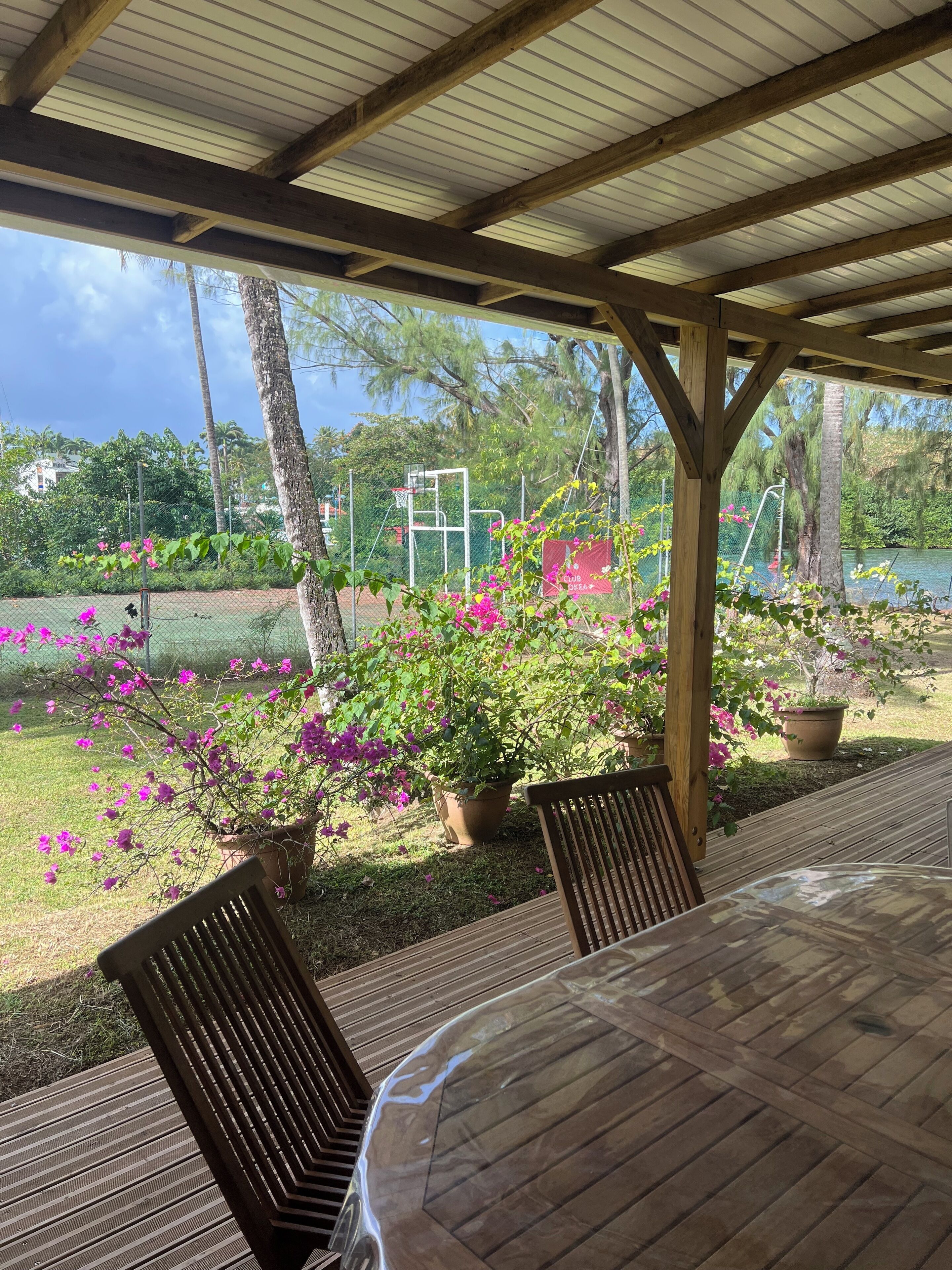 Un patio al aire libre con mobiliario de madera, plantas en macetas y vistas a una cancha de tenis y árboles.