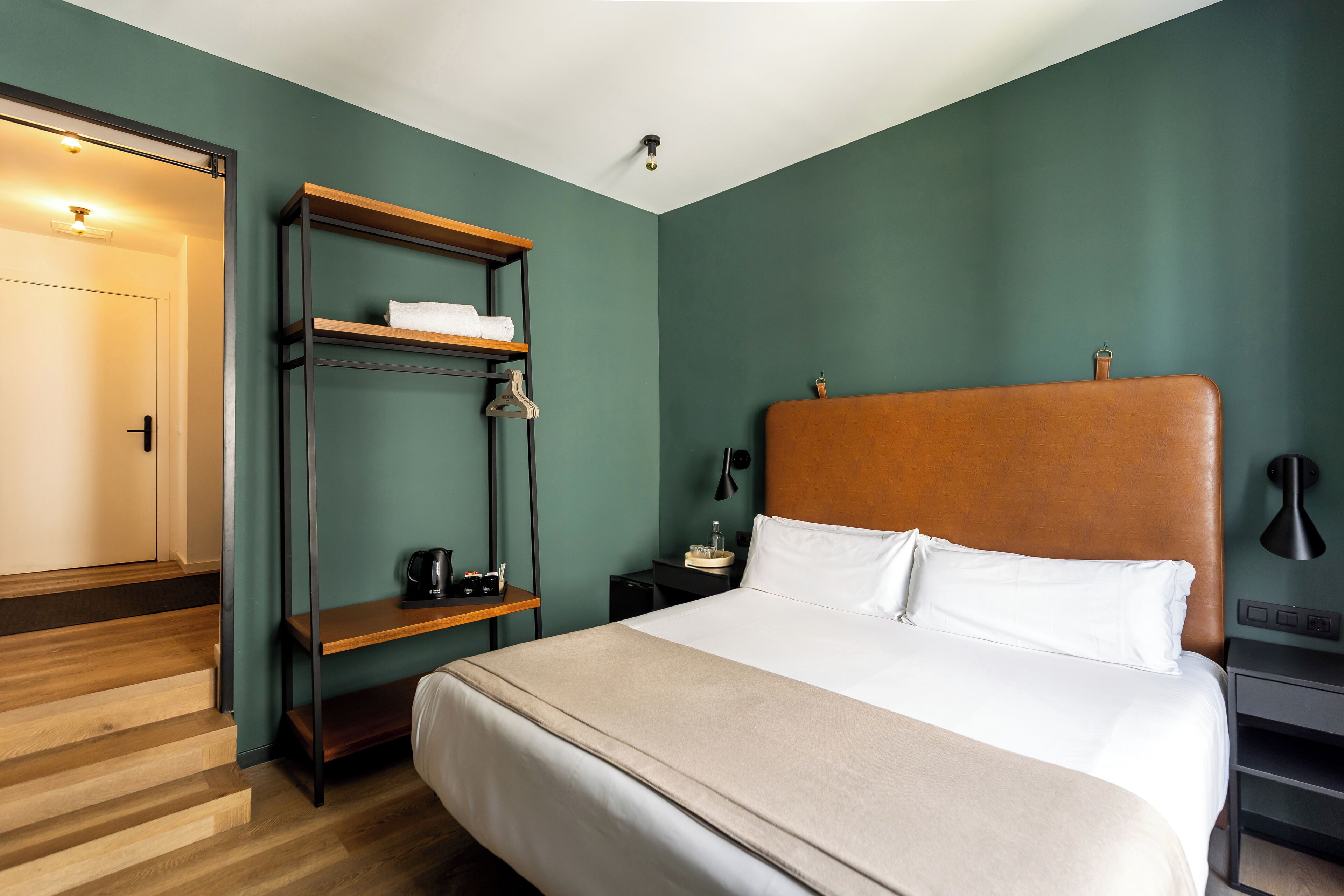 A hotel room with a large bed, a wooden headboard, a shelf unit, a nightstand, and a door.