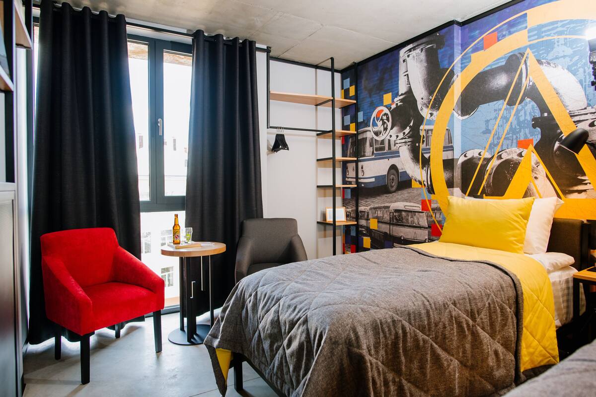 A hotel room with a bed, a red armchair, a small table, and a wall mural.
