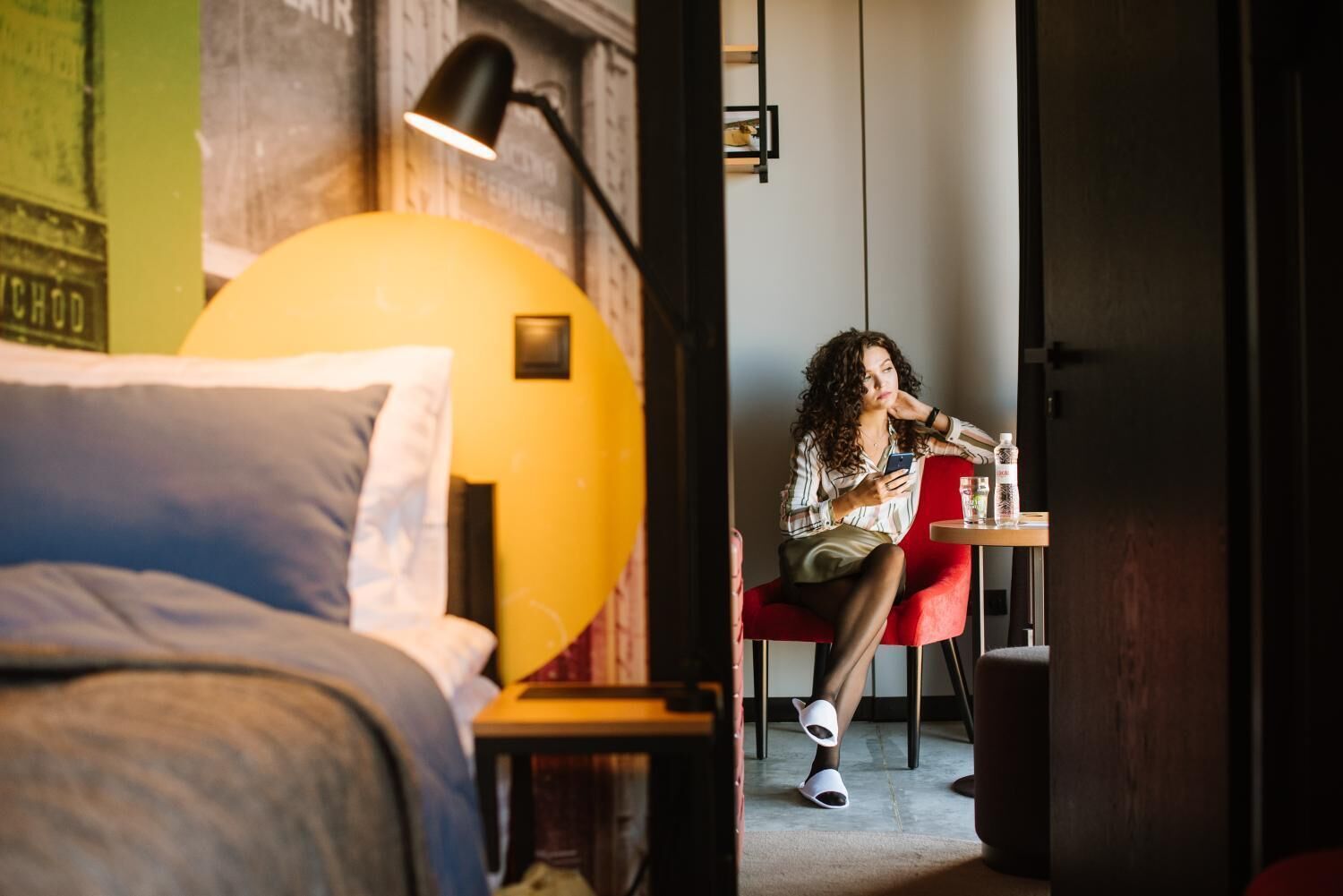 Une pièce avec un lit, une chaise rouge, une table avec une bouteille et une lampe.