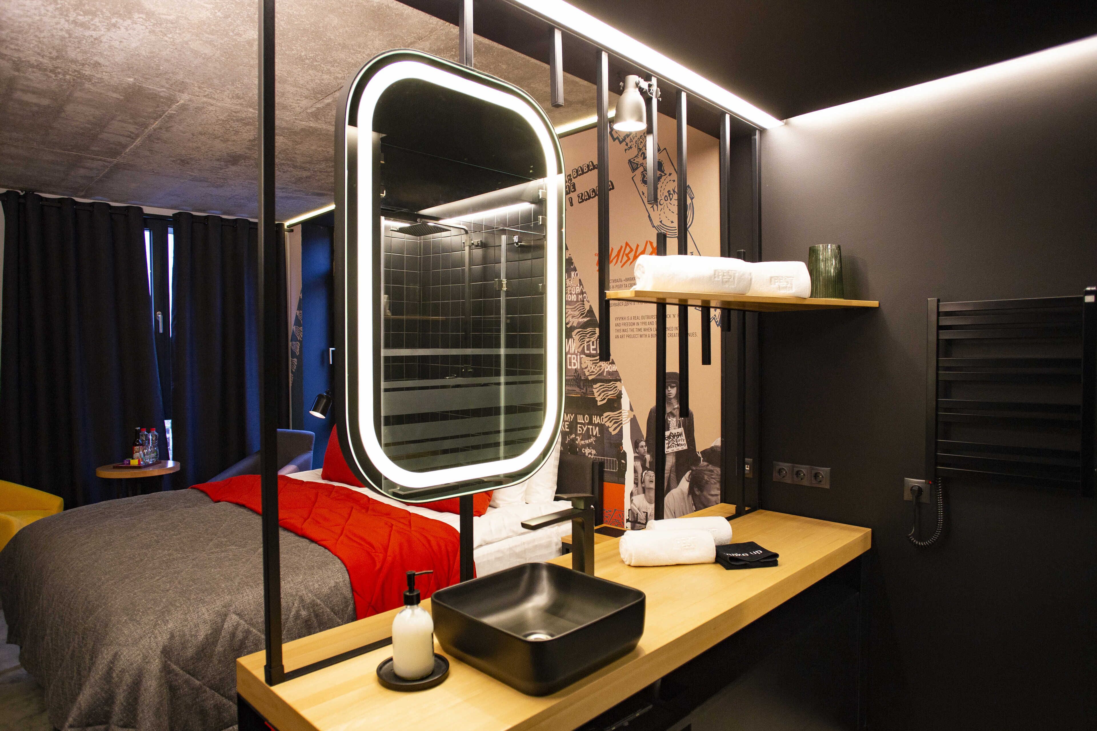 A modern hotel room with a large mirror, a wooden bedside table, a black sink, and a red bedspread.