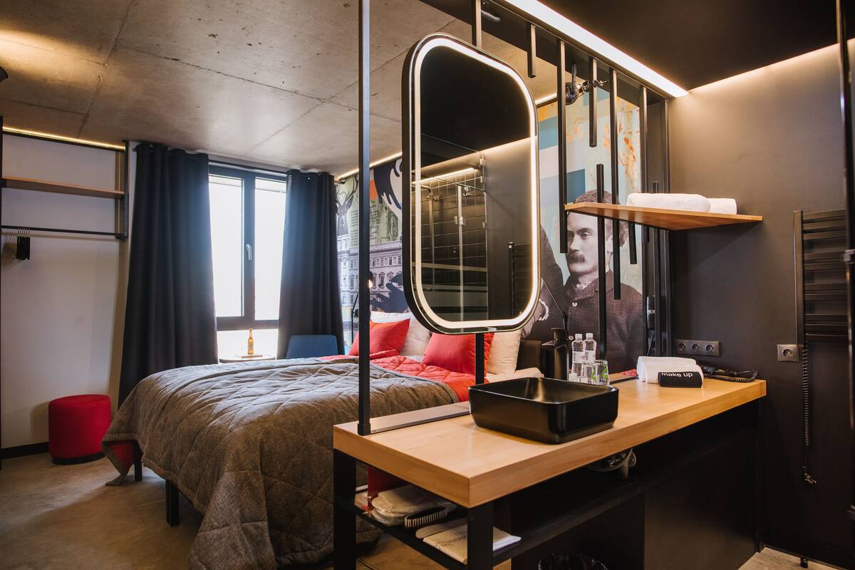 A modern hotel room with a large bed, a vanity with a mirror, a desk with a laptop, and a red ottoman.