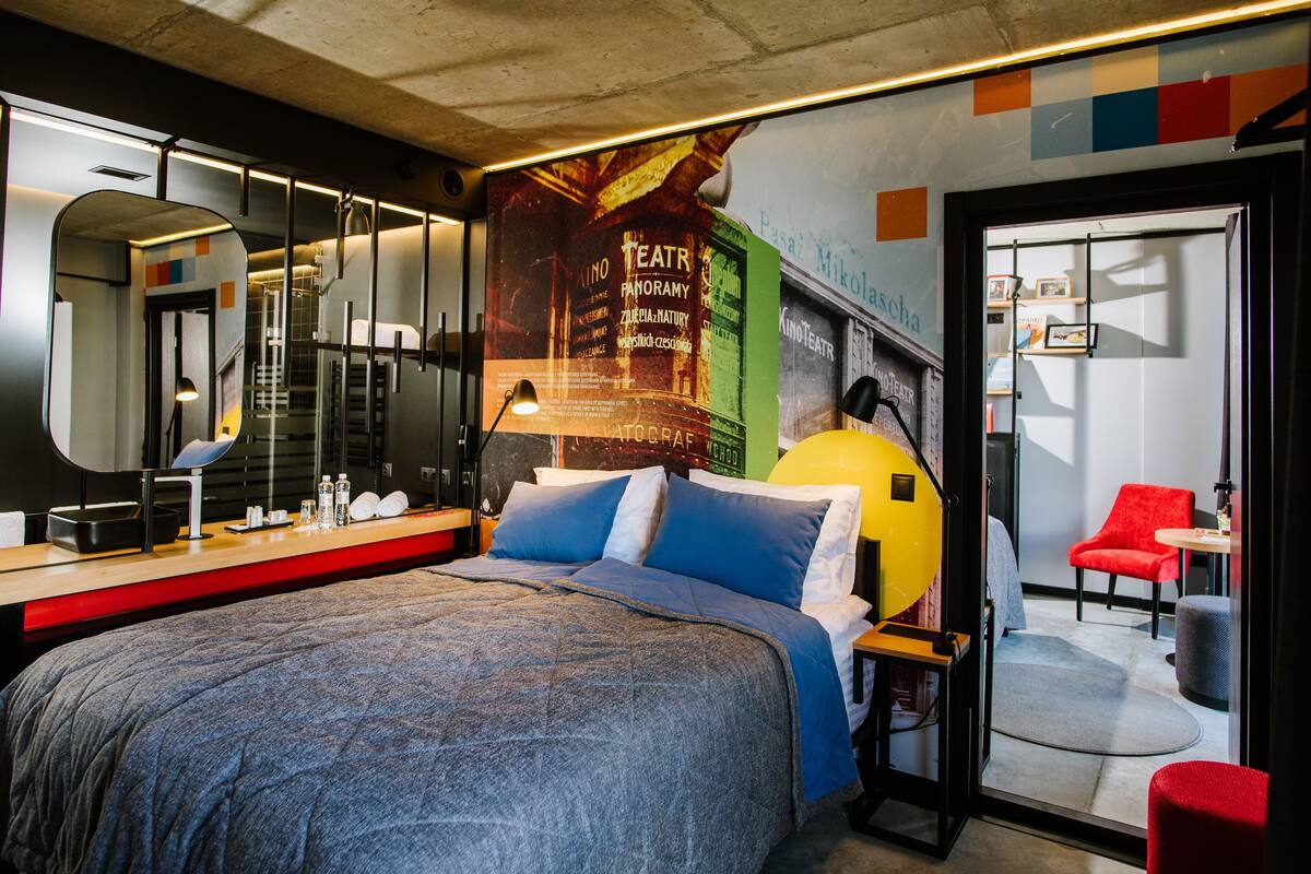 A modern bedroom with a large bed, a mirror, a desk with a lamp, and a red chair.