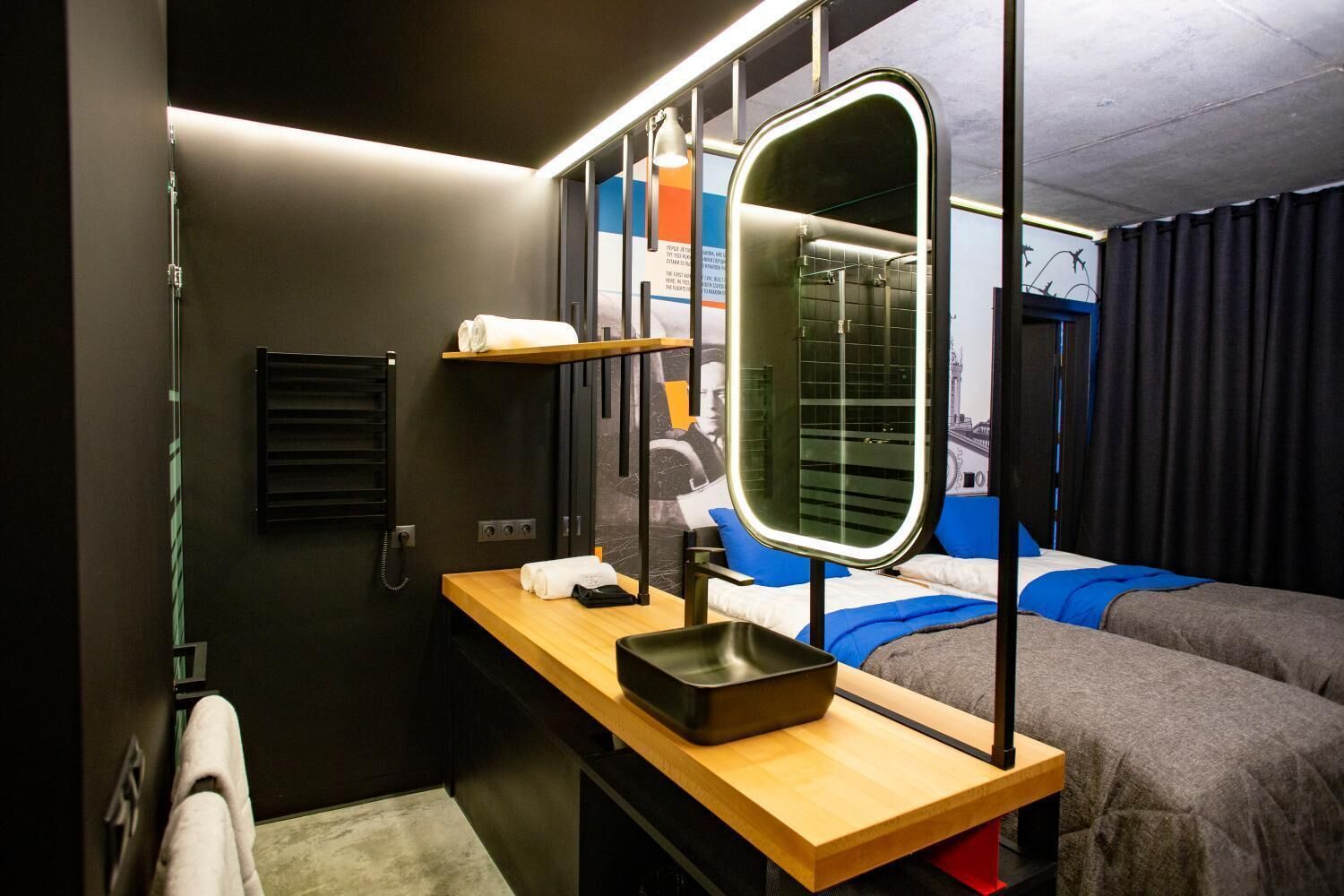 A compact hotel room with a bed, a small table with a mirror, a phone, and a shelf with a towel.