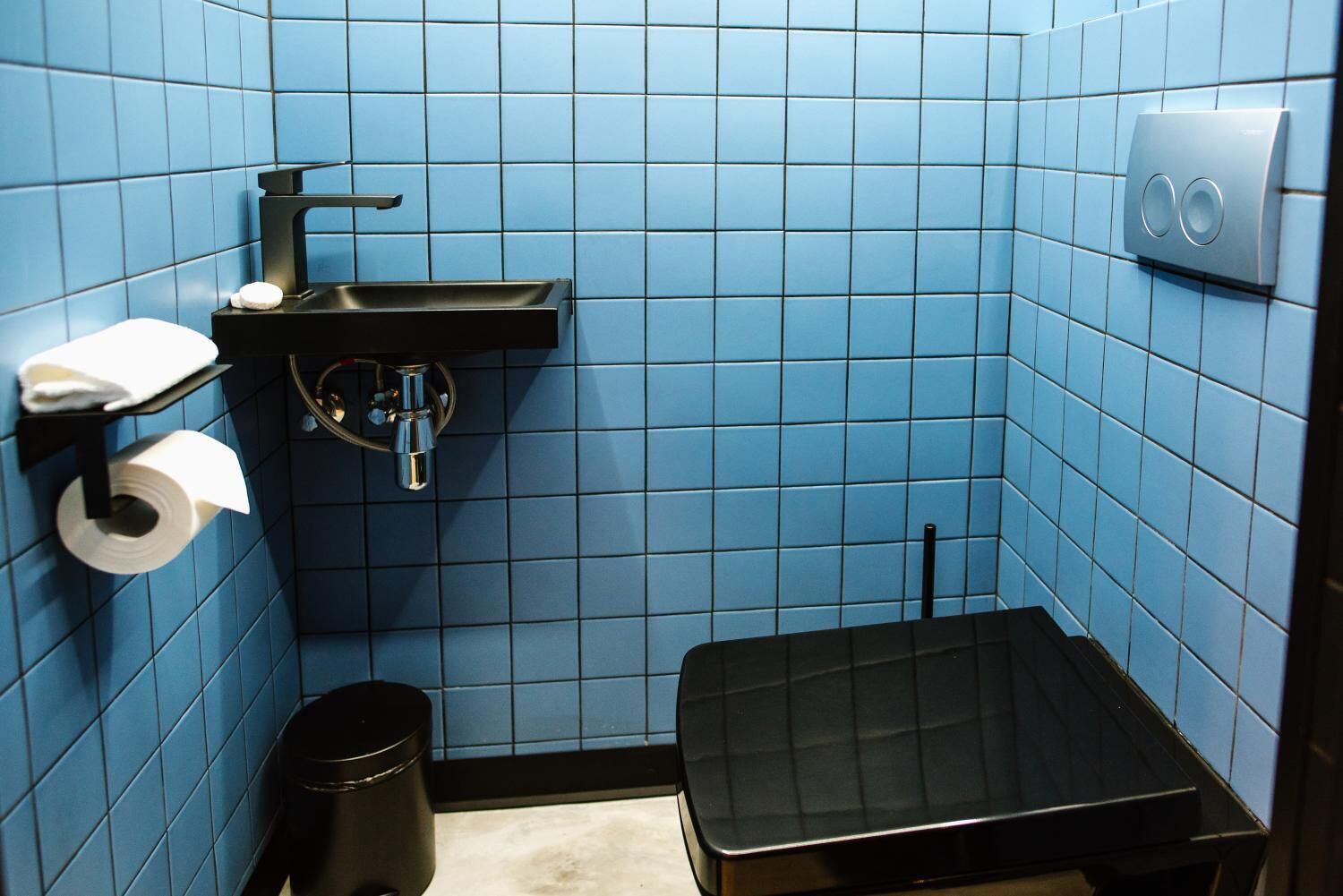 Un baño pequeño con azulejos azules, un lavamanos negro, un dispensador de papel higiénico y un cubo de basura.