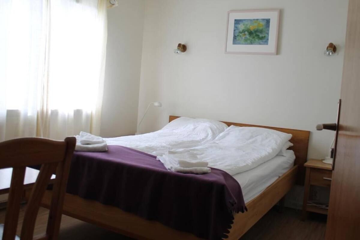 Ein Einzelbett mit weißer Bettwäsche, einer violetten Decke und einem Holz-Kopfteil. Ein kleiner Nachttisch mit Lampe, ein gerahmtes Bild an der Wand und ein Fenster mit durchscheinenden Vorhängen.
