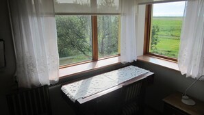 A room with a desk near a window with white curtains, a radiator below, and a lamp on a wooden side table. - Nes Guesthouse (Borgarbyggd)