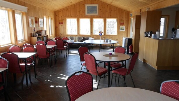 A spacious dining area with multiple tables and chairs, wooden walls, and large windows allowing natural light. - Nes Guesthouse (Borgarbyggd)