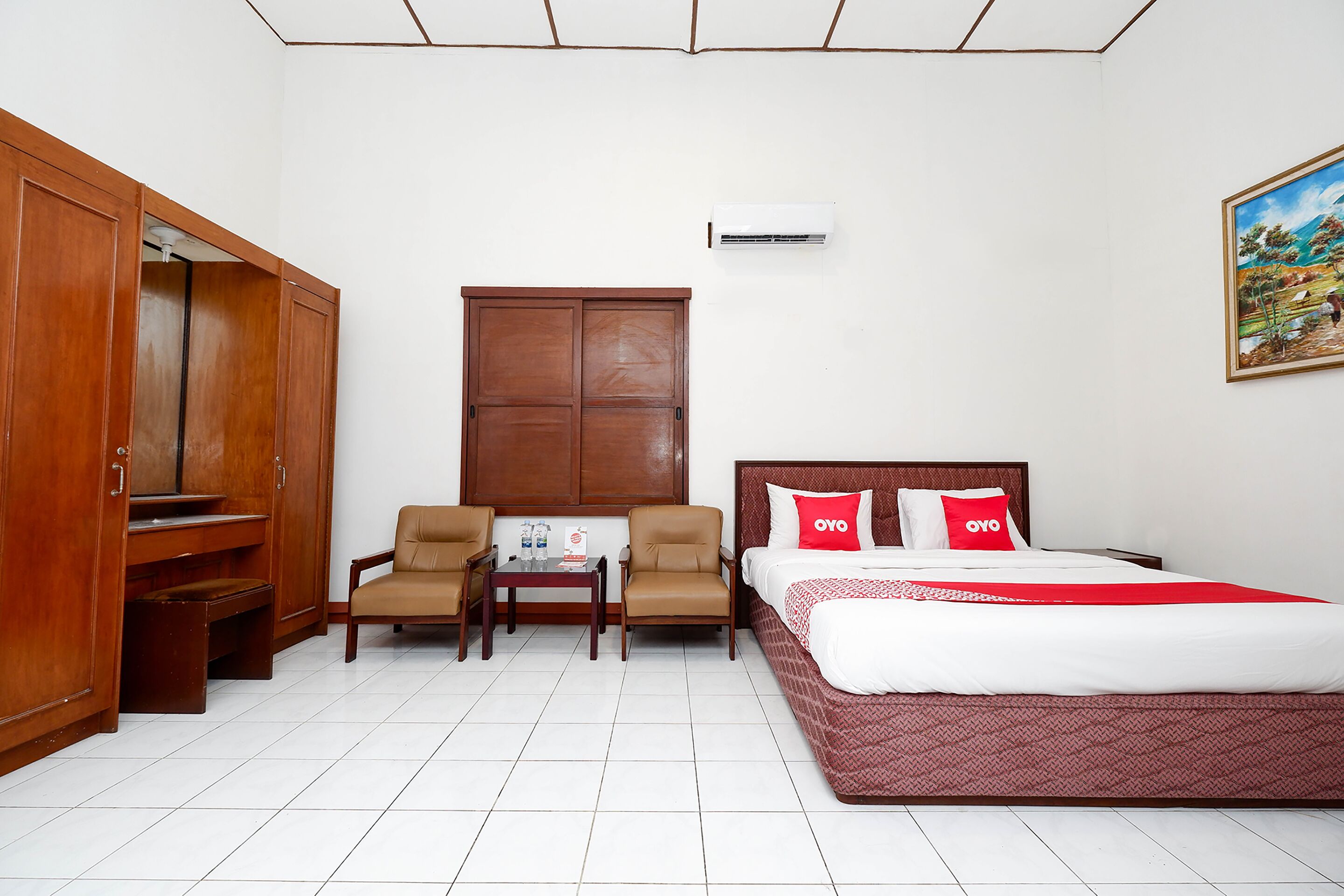 Una habitación de hotel con cama, dos sillas, una mesa, un armario y un cuadro en la pared.