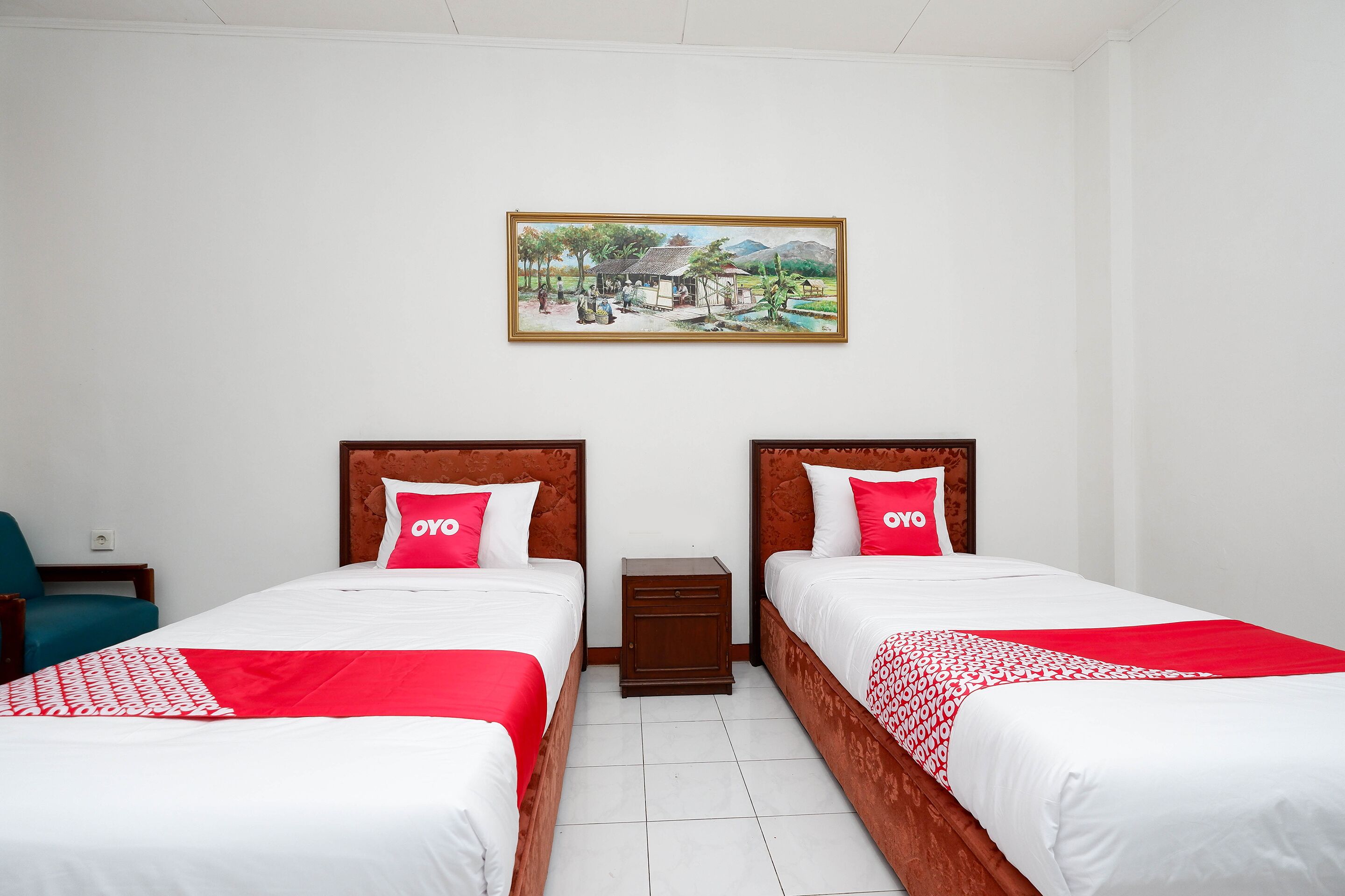 A hotel room with two beds, each with a red and white bedspread and matching pillows. There is a small wooden nightstand between the beds, and a painting hangs on the wall above.