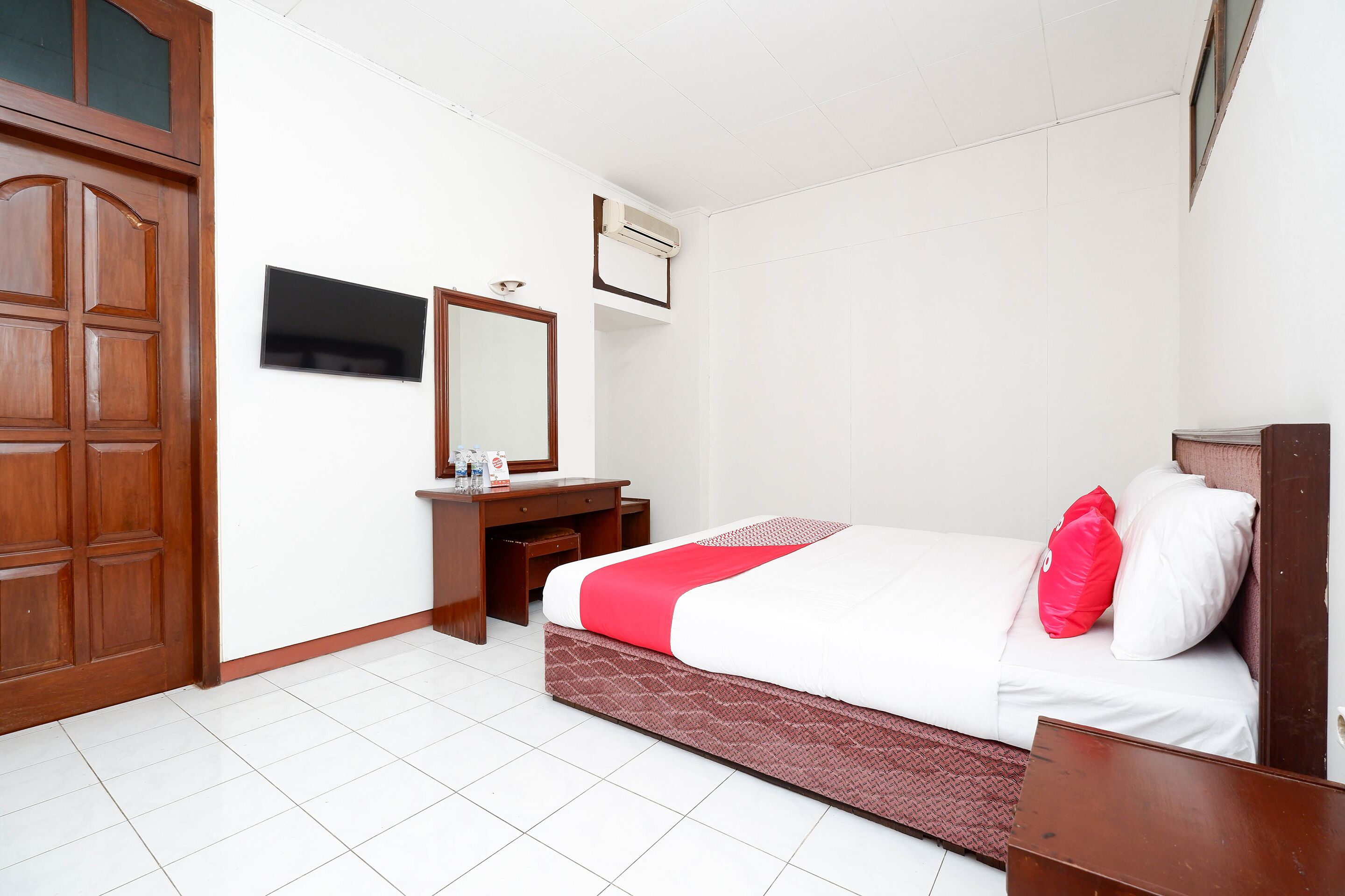 A hotel room with a wooden wardrobe, a desk with a mirror, a bed with white and red bedding, and an air conditioning unit on the wall.