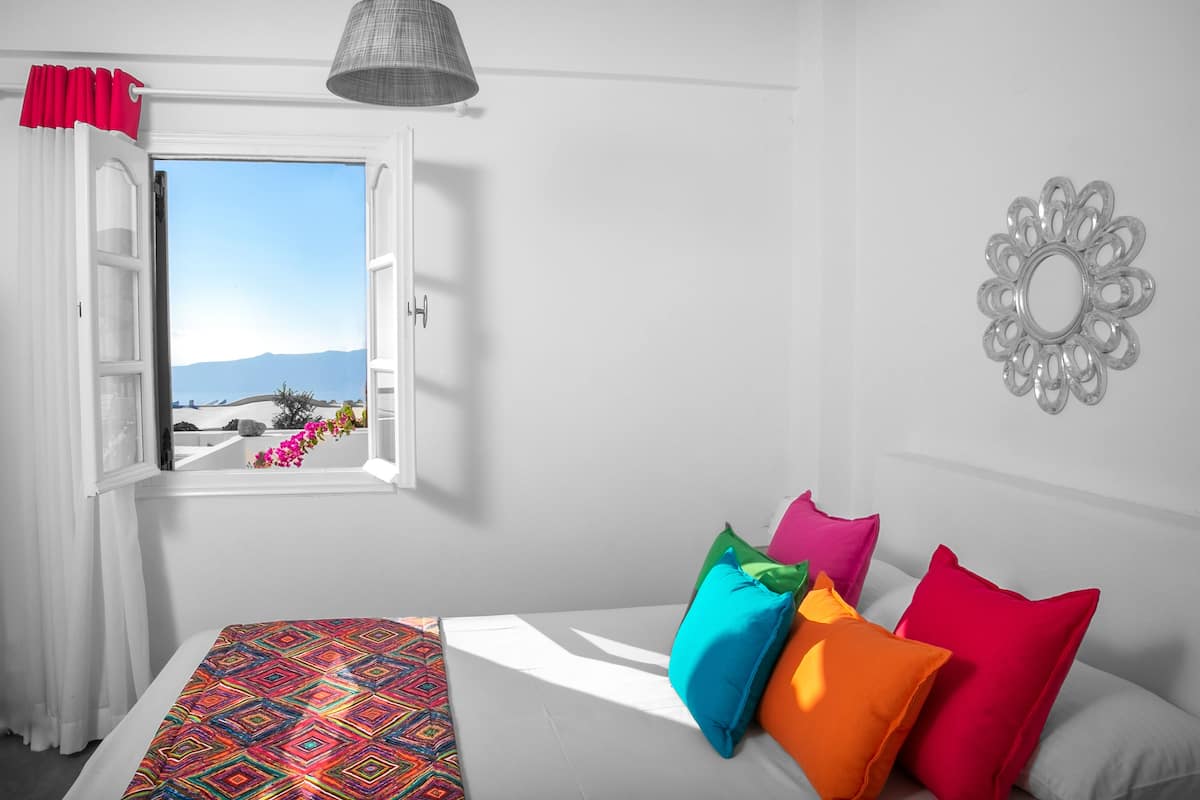 A bedroom with a bed, colorful pillows, a patterned bedspread, a window with curtains, and a wall-mounted mirror.