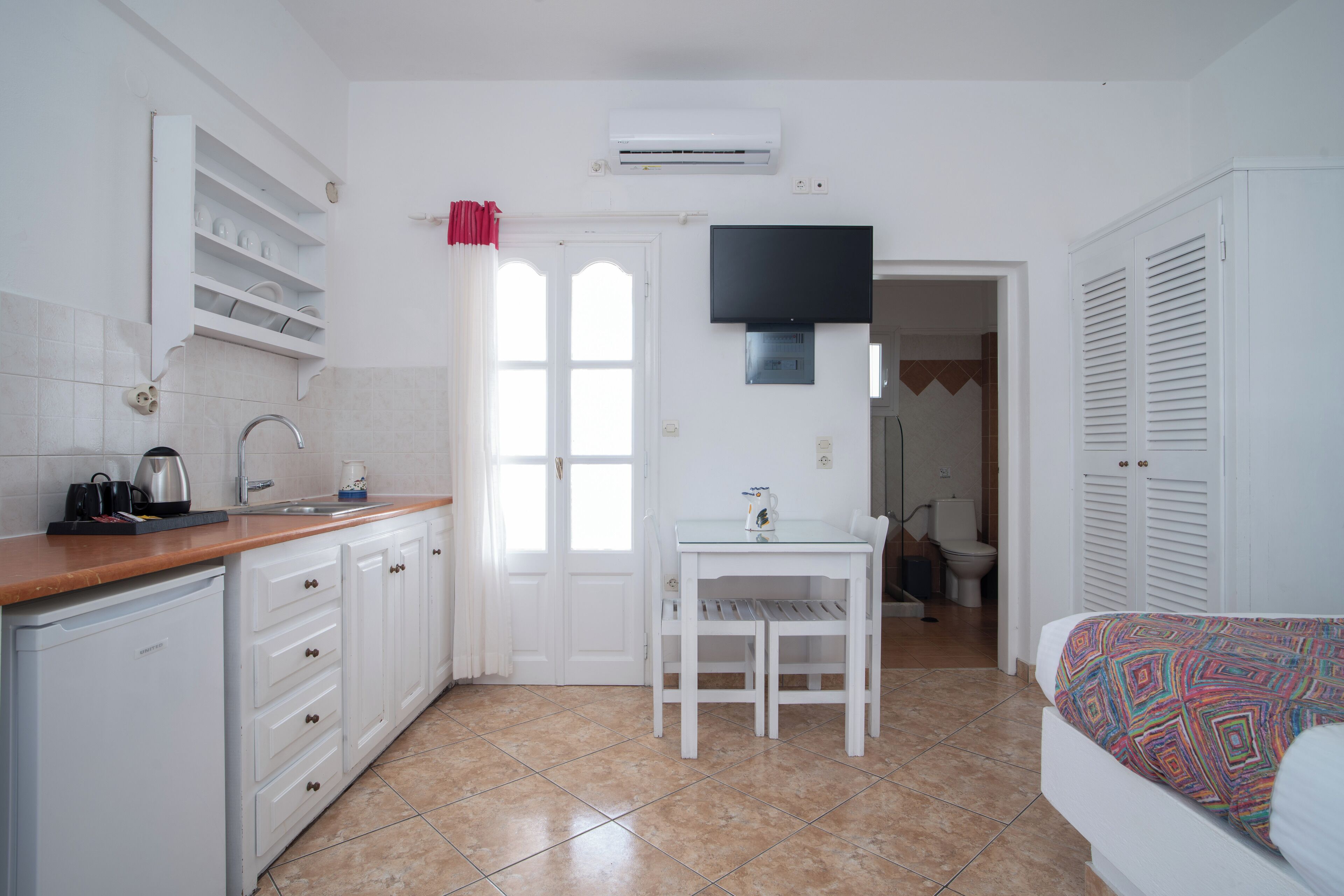 Une cuisine compacte avec des meubles blancs, un évier et un petit réfrigérateur. On y trouve une table à manger avec des chaises et un téléviseur fixé au mur.