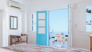A bedroom with a bed, a desk, a chair, a blue door, and a view of the sea. - Salvatore Caldera Suites (Santorini)