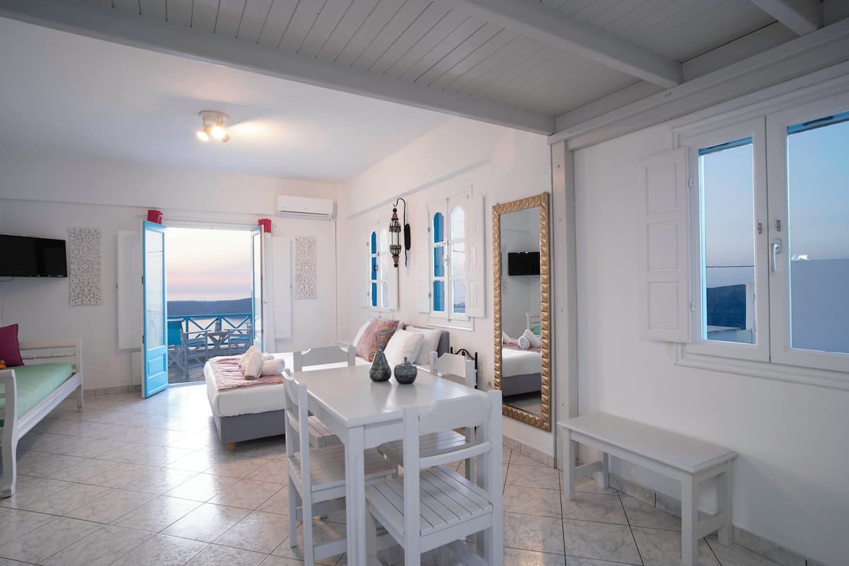 A room with a bed, a dining table, chairs, a bench, a mirror, and a view through an open door.