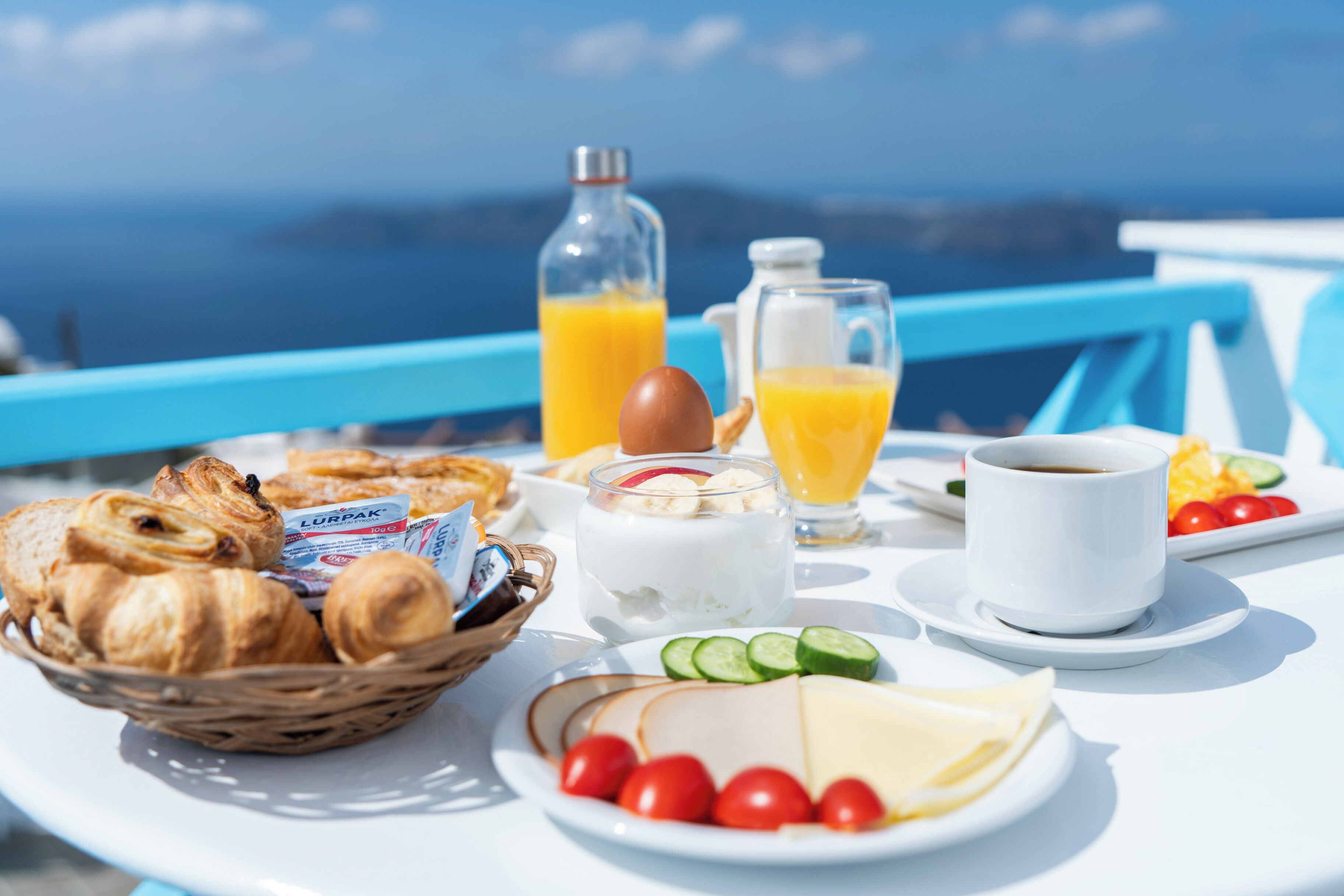 A breakfast table with croissants, cheese, tomatoes, and juice, overlooking a scenic view.