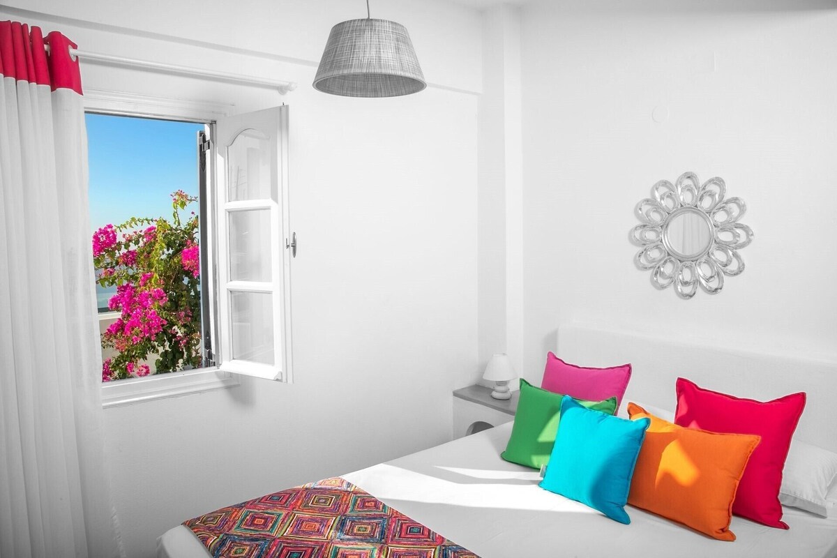 A bedroom with a bed, colorful pillows, a bedside table with a lamp, a window with curtains, and a wall decoration.
