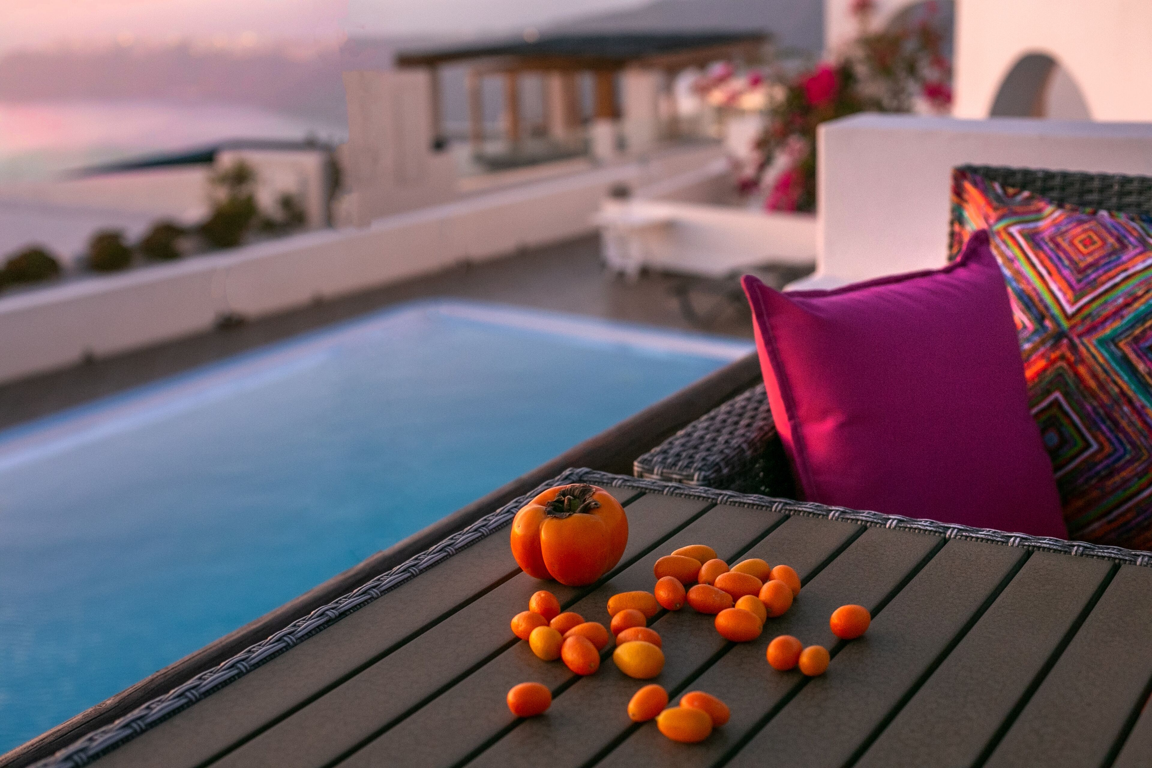 A poolside lounge with a tomato and oranges on a table, a colorful pillow, and a view of the sea.