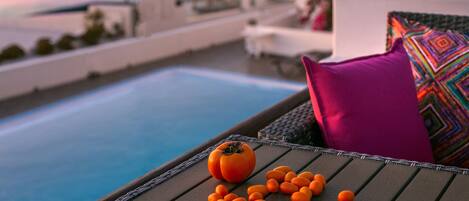 A poolside lounge with a tomato and oranges on a table, a colorful pillow, and a view of the sea.
