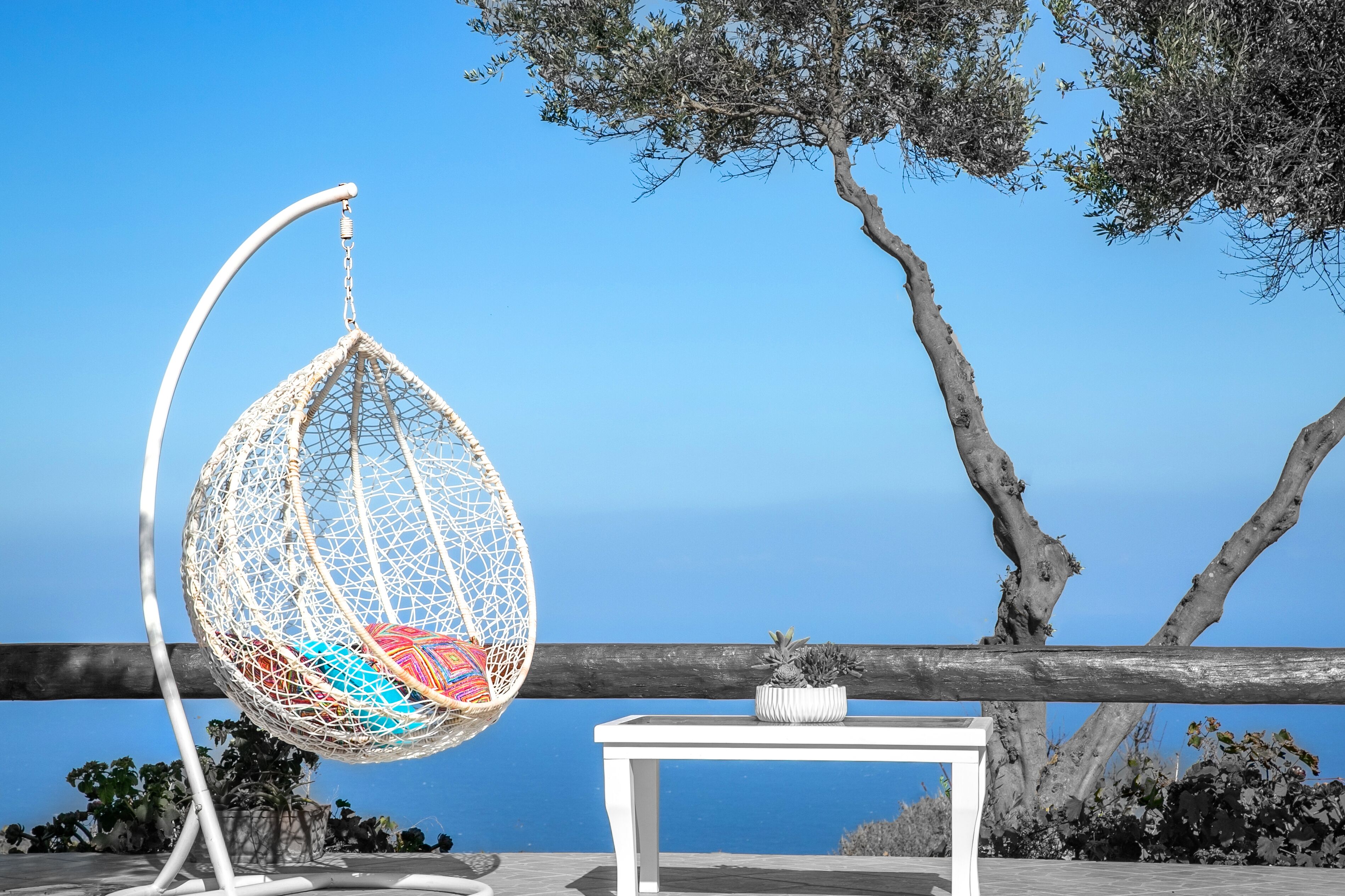 A white hanging chair with colorful cushions, a small white table with a potted plant, and a view of the sea and trees.
