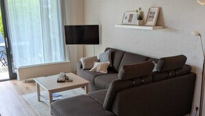 Moderni olohuone, jossa on sohva, sohvapöytä, seinälle asennettu televisio ja ikkuna, jossa on verhot.
