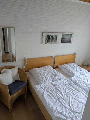 Una habitación con pared de ladrillo blanco, cama de madera con ropa blanca, silla de mimbre con cojines y una mesita con lámpara.