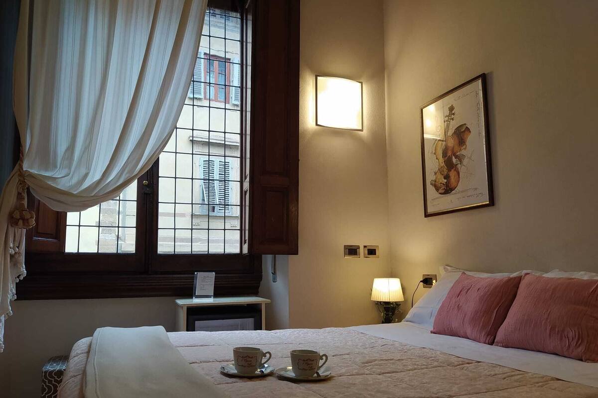 Hotelkamer met een bed, twee bekers op een dienblad, een nachtkastje, een lamp en een raam met gordijnen.