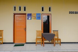 A room with two wooden doors, a small table with a tablecloth, and a chair. There are posters on the wall and a window with a view of trees.